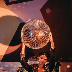 Stylish Woman In Sequined Outfit Holding A Disco Ball At A Lively Party Setting.