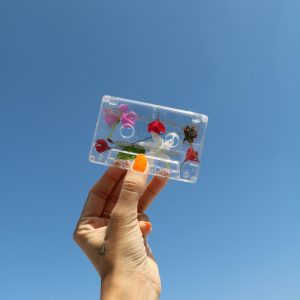 A Hand With Painted Nails Holds A Flower Decorated Cassette Against A Bright Clear Blue Sky, Symbolizing Nostalgia And Creativity.
