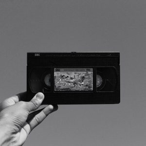 A Hand Holding A Vintage VHS Cassette Tape Against A Plain Gray Background, Evoking Nostalgia.