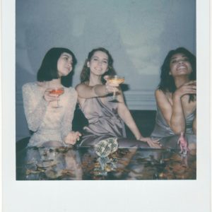 Three Women In Elegant Dresses Enjoying A Celebration With Drinks, Smiling And Having Fun Indoors.