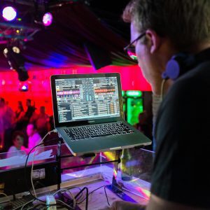 A DJ Plays Music Using A Laptop In A Lively Nightclub With A Vibrant Crowd And Colorful Lighting.