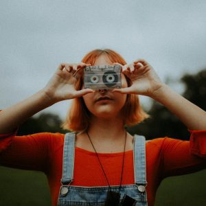 Young Woman With Red Hair Holding A Vintage Cassette Tape, Wearing Bright Red And Denim Under Cloudy Skies.
