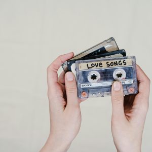 Close Up Of Hands Holding Retro Cassette Tapes Labeled 'Love Songs' On A Light Background.