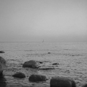 Serene Black And White Seascape With A Distant Sailboat And Rocks In The Foreground.