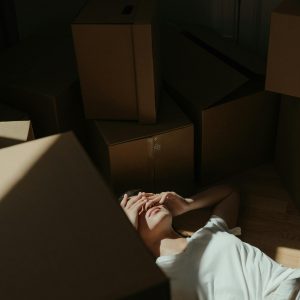 A Woman Lies On The Floor Surrounded By Cardboard Boxes In A Dim Sunlit Room, Expressing Fatigue.