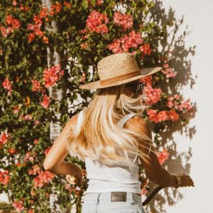 Woman With Long Blonde Hair In A Straw Hat Walking By Blooming Flowers On A Sunny Day. Vibrant And Stylish Lifestyle Scene.