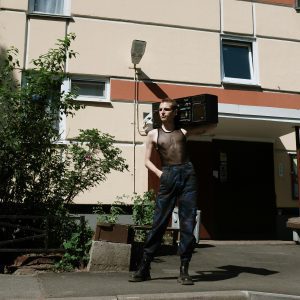A Man In A Mesh Singlet Poses With A Boombox Outside A Panel Building.