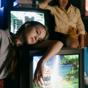 Two Teens Lounging With Vintage CRT Monitors Displaying Video Games In A Dim Room.
