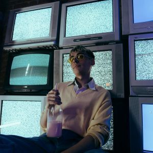 Teenage Boy Holding A Milkshake Surrounded By Vintage CRT Televisions Showing Static In A Dimly Lit Room.