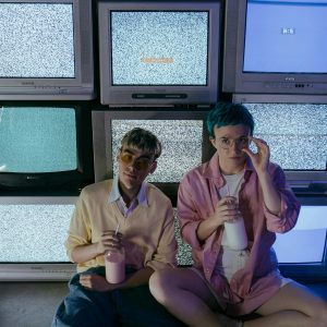 Two Teenagers Enjoying Milkshakes In A Retro Setting With Vintage CRT TVs Displaying Static.