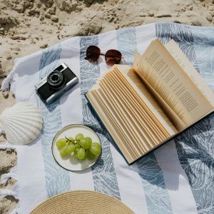 Flat Lay Of Book, Camera, And Grapes On A Beach Towel By The Sand.