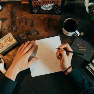 A Top View Of A Vintage Workspace With Drawing Tools, Skull, And Scattered Items On A Rustic Desk.