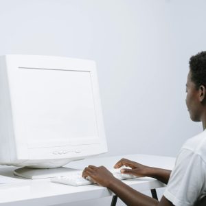 An Adult Man Typing On A White CRT Monitor In A Minimal, Modern Office Setting.