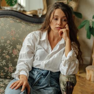 A Woman In A Casual Outfit Sits Thoughtfully On A Floral Vintage Sofa Indoors.
