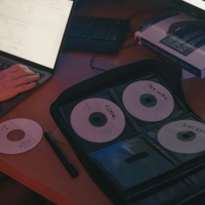 A Person Works On A Laptop Beside A Set Of Labeled CDs On A Desk.