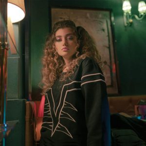 Low Angle Of Young Curly Haired Female With Bright Makeup In Casual Clothes Standing Near Mirror At Home And Looking At Camera