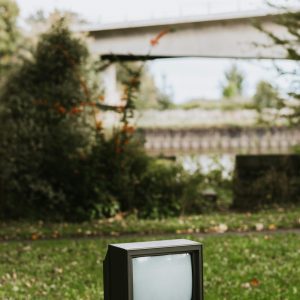 Small Black Old Fashioned Television Placed On Bench In City On Street With Green Plant And Bridge On Blurred Background