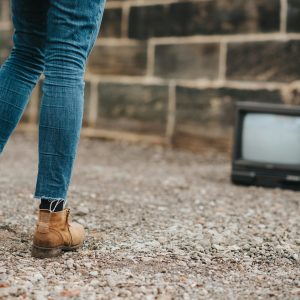 Back View Of Anonymous Female In Jeans Standing On Ground Against Stone Wall And Old Fashioned Television On Blurred Background