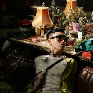 A Fashionable Individual Poses In A Vintage Setting Adorned With Eclectic Accessories And Warm Lighting.