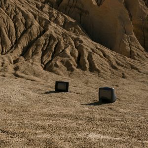 Two Vintage Televisions On A Barren Desert Landscape Against Rugged Hills Under A Blue Sky.