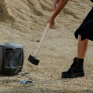 A Person In Black Boots And Dress Smashes An Old Television With A Mallet On Sandy Ground.
