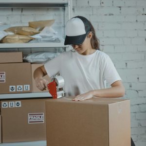 Young Female Worker Sealing Cardboard Boxes In An Indoor Warehouse Setting.