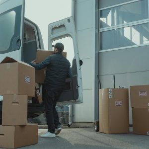 A Delivery Man Unloading Cardboard Boxes From A Van At A Warehouse During The Day.