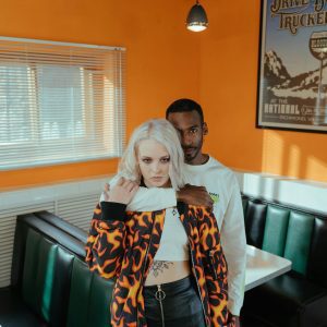 A Loving Multiracial Couple Embraces In A Retro Diner With Vibrant Orange Walls.