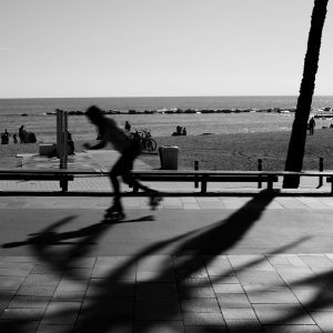 A Silhouette Of A Roller Skater Swiftly Moves On A Beachfront Boardwalk In A Black And White Scene.