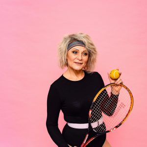 Elderly Woman Posing With Tennis Equipment Against A Pink Backdrop.