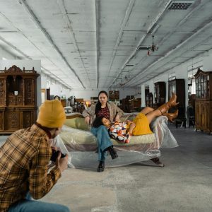 Model Posing On Vintage Furniture With Photographer Capturing The Scene In Atmospheric Warehouse.
