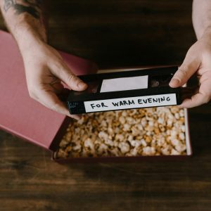 Hands Holding A Vintage VHS Tape Over A Box Of Popcorn On A Wooden Surface.