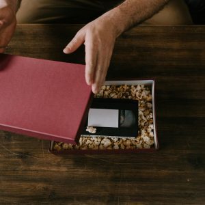 Hands Opening A Maroon Gift Box Containing A VHS Tape Cushioned In Popcorn.