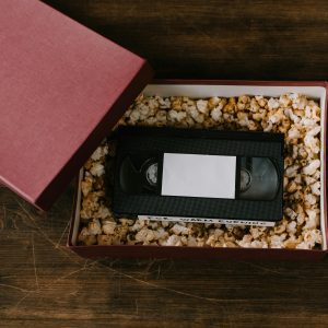 An Open Gift Box With A VHS Tape Nestled In Popcorn, On A Wooden Surface.