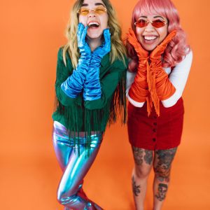 Two Women In Colorful Outfits And Wigs Express Joy Against An Orange Backdrop.