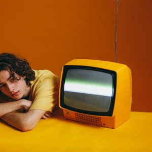 A Young Man With Curly Hair Rests Beside A Vintage Orange TV On A Vibrant Backdrop.