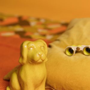 A Vibrant Yellow Ceramic Dog Figurine On An Orange Bed With Binoculars And Colorful Patterns In The Background.