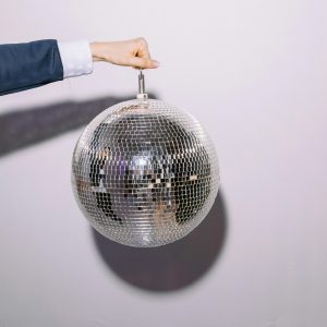 A Hand Holding A Reflective Disco Ball Against A Plain White Background, Casting A Shadow.