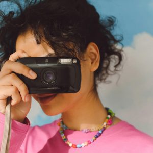 A Young Woman With Curly Hair Holds A Retro Camera, Standing Against A Sky Background With Clouds.