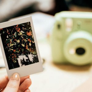 Hand Holding An Instax Photo Of Orange Berries With Blurred Instant Camera In The Background.