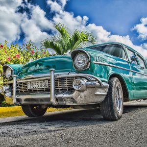 A Beautifully Restored Classic Car On A Sunny Road In Havana, Cuba, With Lush Foliage And A Vibrant Sky.