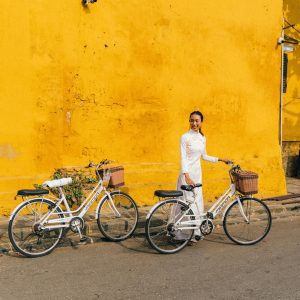 Vietnamese Woman In Traditional Dress With Bicycles In Hoi An, Vietnam Against A Yellow Wall.