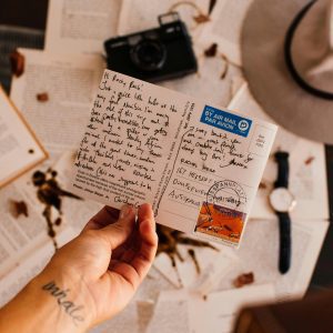 A Hand Holding A Handwritten Postcard Surrounded By Travel Essentials On A Table.