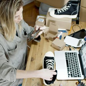 Woman Photographing Shoes For Online Sale In Home Workspace With Laptop And Packages.