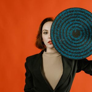 A Stylish Woman Partially Covers Her Face With A Round Artistic Disk Featuring Script On A Striking Red Background.