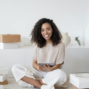 A Confident Young Woman Sitting Among Packages And Papers While Using A Tablet, Showcasing Modern Entrepreneurship.