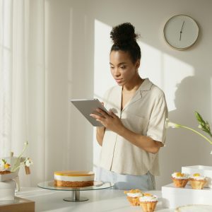 A Woman Using A Tablet In A Bright And Modern Kitchen Setting, Surrounded By Cakes And Flowers, Depicting A Lifestyle Scene.