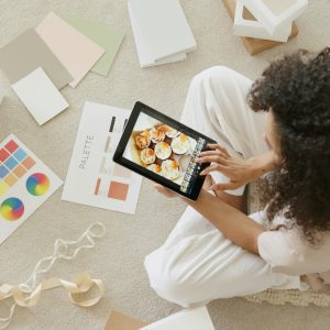 Woman Using Tablet Surrounded By Design Tools And Gift Box, Showcasing Creativity And Online Work.