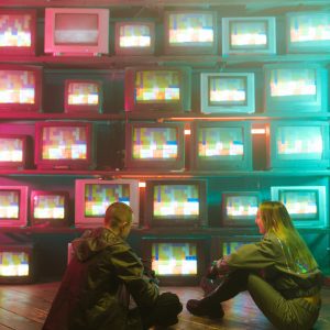 Two Gamers In Futuristic Attire Sit By A Neon Lit Wall Of Retro TVs.
