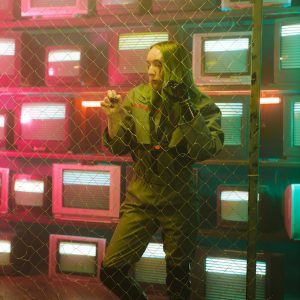 A Woman With A Prosthetic Hand Stands Against A Chain Link Fence With Colorful Retro Tv Screens.